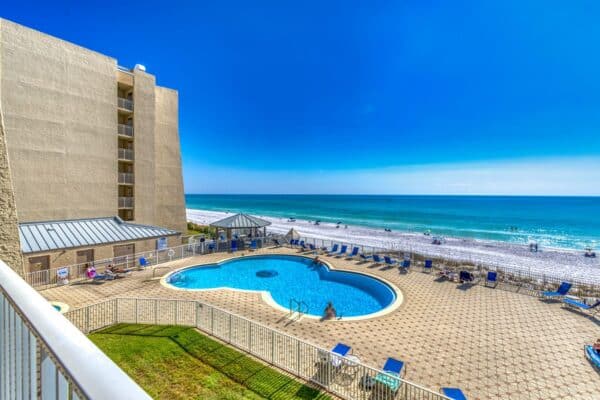 A beachfront hotel with a large, circular swimming pool surrounded by lounge chairs, overlooking the ocean on a sunny day. For those seeking a room close to all amenities, C303 offers convenience and breathtaking views.