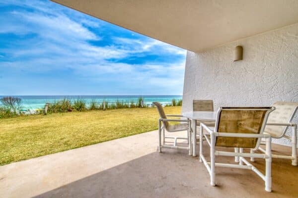 A covered patio with a table and four chairs, marked as D104, overlooking grassy area and ocean under partly cloudy sky.