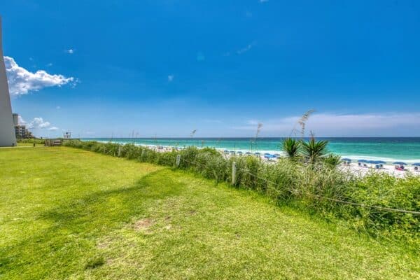 A grassy path leads to a sandy beach with rows of blue umbrellas by the ocean under a clear blue sky at B101.