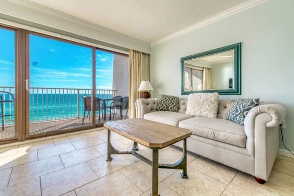 Living room with a light sofa, wooden coffee table, and sliding glass door opening to a balcony with ocean view. Mirror on wall.