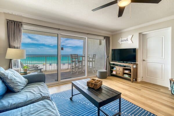 Living room with blue sofa, wooden coffee table, TV, and balcony view of beach and ocean at a condominium complex.