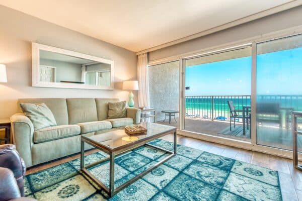 A living room with a beige couch, glass coffee table, large mirror, and ocean view through sliding glass doors. A patterned blue rug on the wooden floor.