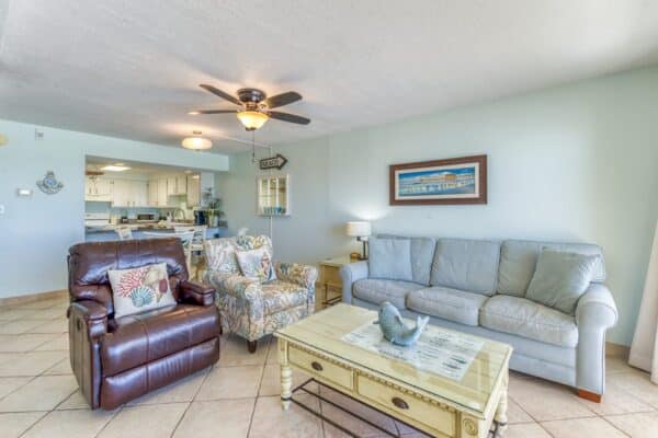 Living room with a ceiling fan, leather recliner, floral armchair, light gray sofa, coffee table with bird figurine, and kitchen in the background.