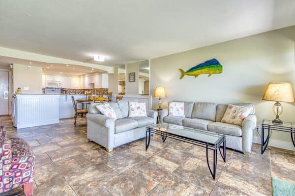 Bright living room with a modern kitchen at The Beach House Condos, furnished with a plush sofa, glass tables and fish wall decor.