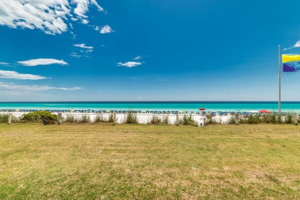 Coastal scene at The Beach House Condominiums with a grassy area leading to a beach lined with colorful umbrellas under a clear sky.