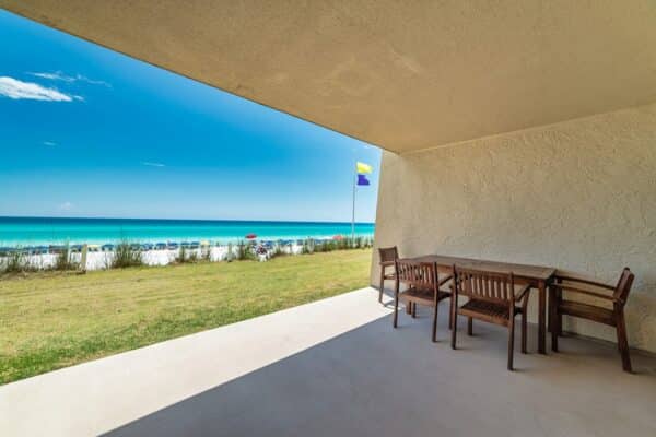 Beachfront patio with a wooden dining set at The Beach House Condominiums, overlooking a calm sea under a clear sky, featuring D102.