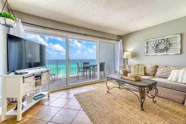 A living room with a large TV, beige sectional sofa, and coffee table. Glass doors open to a balcony with an ocean view.