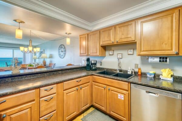 Kitchen with wood cabinets, black counters, stainless dishwasher, sink, coffee maker, and view of dining area with chandelier.