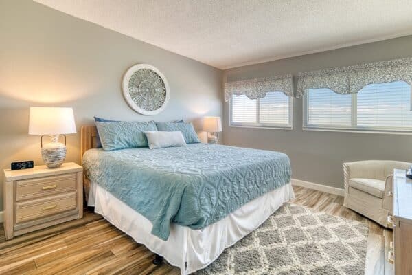 Neatly made bed with blue quilt, pillows, two lamps, three windows, armchair, and patterned rug in a bright condo bedroom.