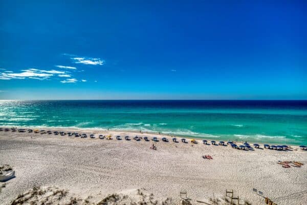 Sandy beach with lounge chairs and umbrellas facing turquoise ocean, clear sky; view from a condominium complex.