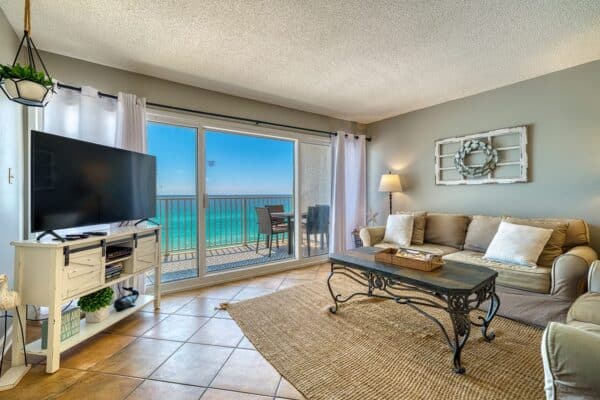 A living room with sofa, coffee table, TV, and sliding doors to a balcony with ocean view; tiled floors and neutral decor.