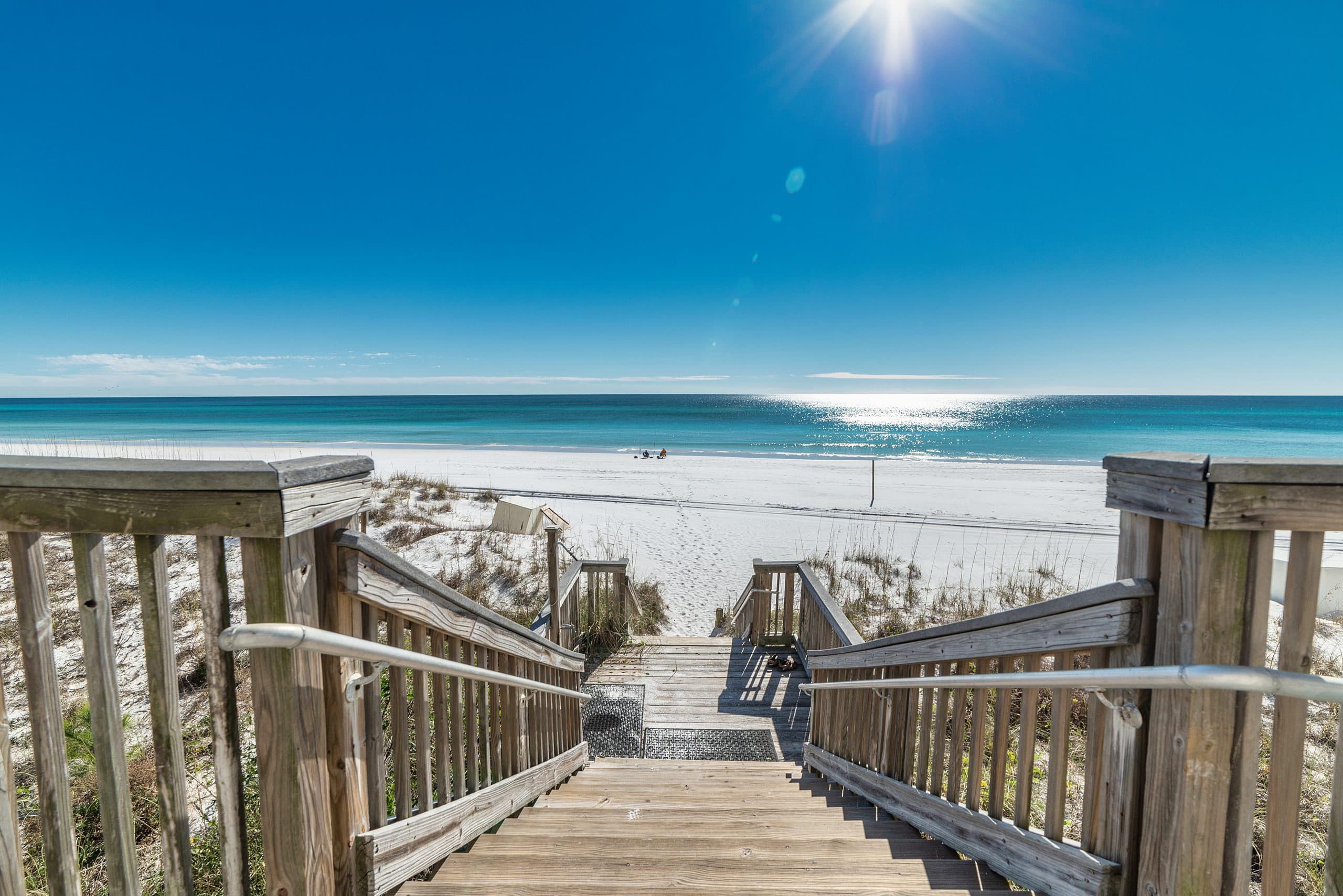 Wooden boardwalk at The Beach House Condominiums leading to a sunny beachfront with clear blue skies.