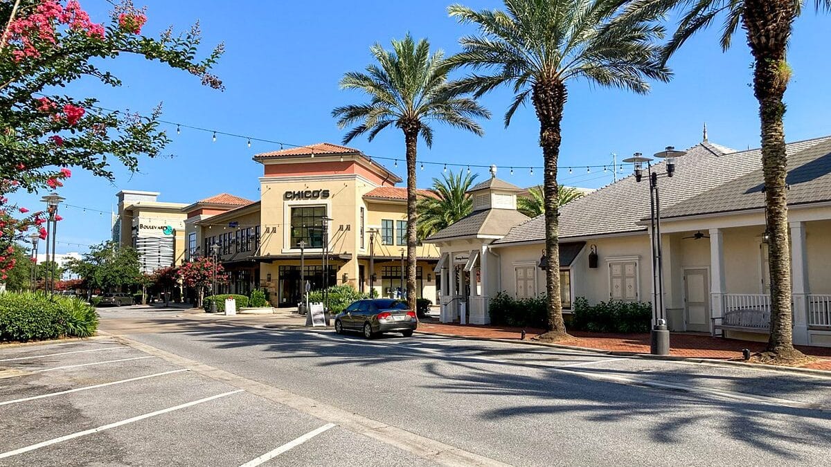 Sunny street view of a shopping area near The Beach House Condominiums with palm trees and a Chico's store.