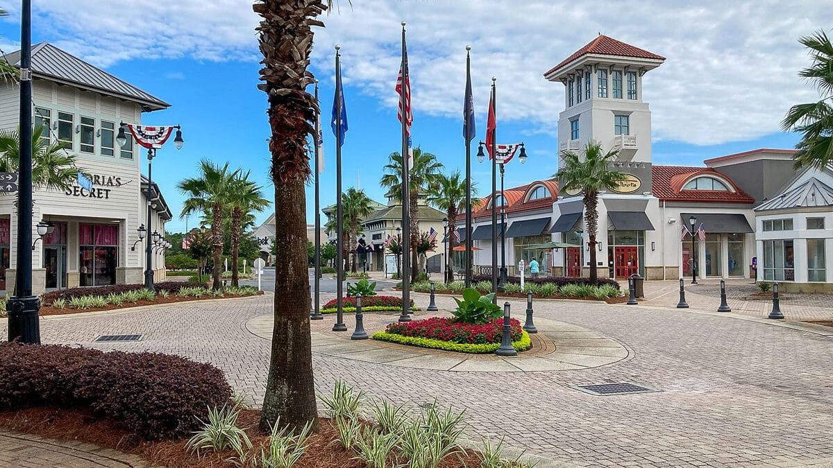 Pedestrian zone in The Beach House Condominiums' shopping district, featuring palm trees, American flags and storefronts on a partly cloudy day.