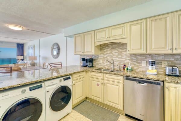Modern kitchen with granite counter, light cabinets, appliances, and stacked washer/dryer; dining and living areas behind.