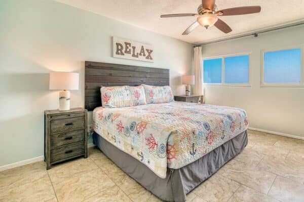 Bedroom with king bed, wood headboard, nightstands, lamps, ceiling fan, "RELAX" sign, light walls, and tile floor.