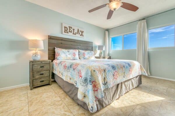 A bedroom with a bed and colorful quilt, two bedside tables, a ceiling fan, and a window. A "RELAX" sign is above the headboard.