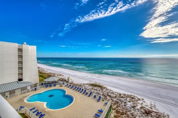 View of a beachfront with a heart-shaped pool and blue lounge chairs next to a white building under a blue sky.