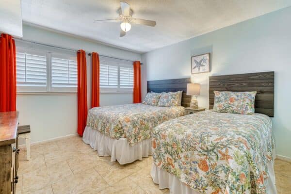 Bright bedroom at the condominium with two double beds, floral bedding, red curtains, tiled floor, and beachy wall art.