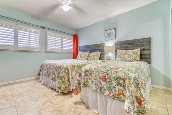 Two beds with floral bedspreads, a ceiling fan, lamp, and windows with red curtains in a light blue bedroom.