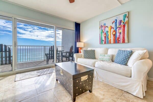 Living room with a white sofa, decorative pillows, and wooden coffee table; large windows show ocean view. Parrot painting on wall.