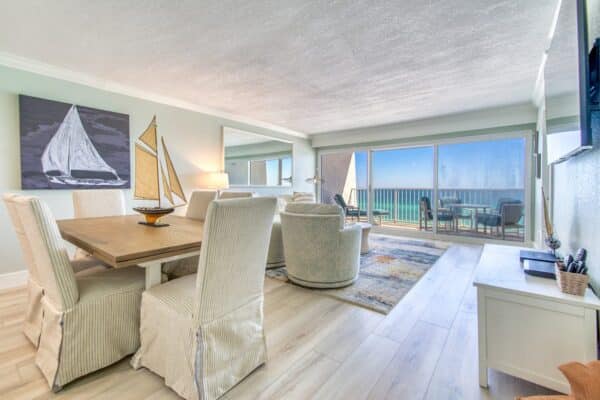 Modern beachfront living room in The Beach House Condominiums with a dining table, plush chairs, nautical decor, and ocean views through sliding glass doors.