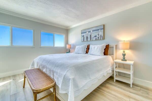 Brightly lit bedroom at Beach House Condominiums, featuring white bedspread, wooden bed set, side tables with lamps and a cityscape art piece.