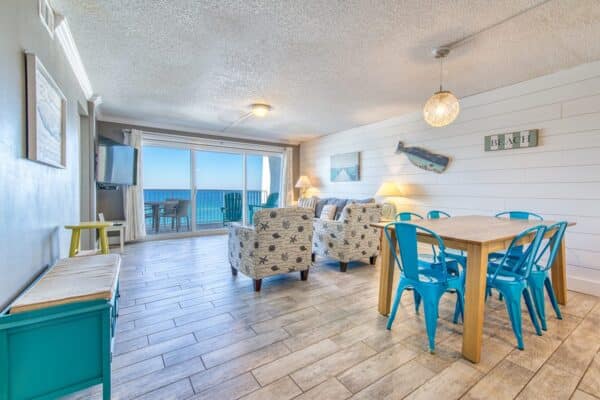 B502 living and dining area with blue chairs, wooden dining table, patterned armchairs, large ocean-view window, and beach-themed decor.