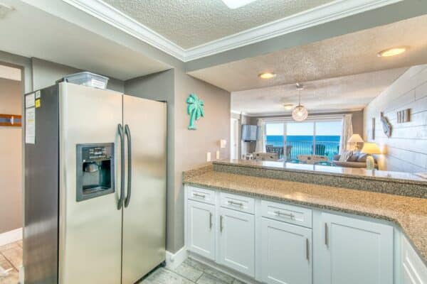 Modern kitchen with stainless steel refrigerator, granite countertops, and white cabinets. Dining area with beachfront view in the background.