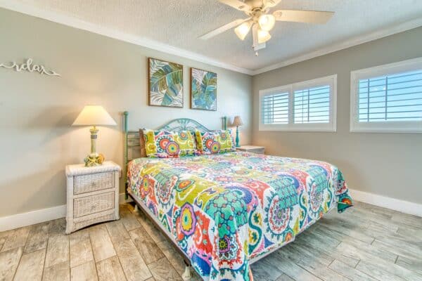 A bedroom with a colorful quilt on the bed, two framed artworks above the headboard, a ceiling fan, and windows with white shutters.