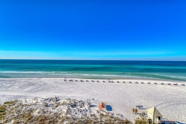 A sandy beach with rows of lounge chairs and umbrellas faces a calm, blue ocean under a clear sky.