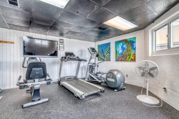 Compact gym at The Beach House Condos with treadmill, bike, elliptical, TV, wall art and a fan.