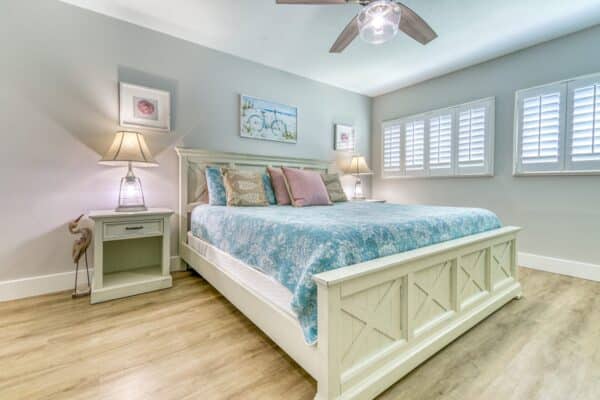 A neatly arranged bedroom with a queen-sized bed, blue patterned bedspread, two nightstands with lamps, ceiling fan, and white plantation shutters.