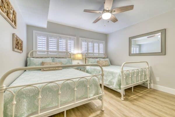 A bedroom with two white metal frame beds with green bedding, a ceiling fan, light-colored walls, and wooden floors.