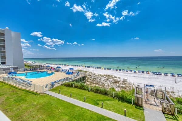 A beachfront with umbrellas, lounge chairs by the shore, a fenced pool area, and Building C301 under a clear blue sky.